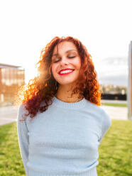 Cheerful young female in blue sweater with curly red hair holding hands behind back and smiling with closed eyes while standing near grassy lawn at sunrise in city - ADSF42699