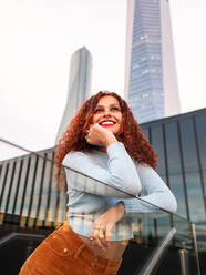 Fröhliche junge Frau mit langem lockigem rotem Haar und Lippen im Pullover, die sich an einen Glaszaun lehnt und in der Nähe eines modernen Wolkenkratzers in der Stadt wegschaut - ADSF42703