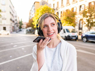 Positive junge Frau mit blonden Haaren in Freizeitkleidung und kabellosen Kopfhörern, die lächelnd eine Sprachnachricht über ihr Smartphone aufnimmt, während sie an einem sonnigen Herbsttag auf einer Straße in der Stadt steht - ADSF42706