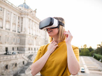 Low angle of positive young female with blond hair in yellow t shirt and VR goggles smiling while watching video and exploring virtual reality near historic palace in city - ADSF42708