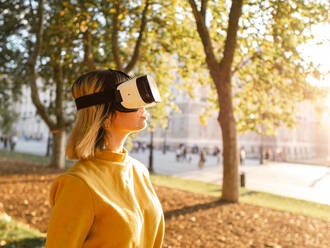 Serious young female with blond hair in yellow t shirt and VR goggles watching video and exploring virtual reality in park - ADSF42709