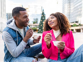 Fröhlicher junger hispanischer Freund und afroamerikanische Freundin sitzen auf einer Bank in der Stadt und lachen zusammen, während sie Eis aus Plastikbechern essen - ADSF42711