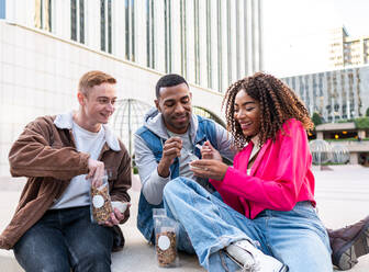 Eine Gruppe multiethnischer Studenten in Freizeitkleidung sitzt in der Nähe eines Universitätsgebäudes und isst leckere Snacks, während sie miteinander plaudern - ADSF42712