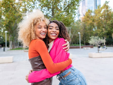 Lächelnde junge Studentinnen in farbenfrohen, modischen Outfits mit lockigem Haar, die in einem städtischen Park stehen und sich umarmen, während sie sich gegenseitig ansehen - ADSF42714