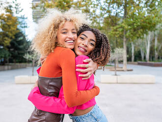 Lächelnde junge Studentinnen in farbenfrohen, modischen Outfits mit lockigem Haar, die in einem städtischen Park stehen und sich umarmen, während sie sich gegenseitig ansehen - ADSF42715