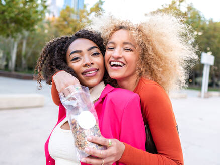 Fröhliche multirassische Freundinnen mit lockigem Haar, die sich auf der Straße umarmen und Nüsse aus einer Plastikverpackung auf einer Stadtstraße mit unscharfem Hintergrund essen und in die Kamera schauen - ADSF42717