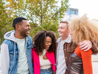 Gruppe von fröhlichen multirassischen Freunden in Streetstyle-Outfits, die im Park in der Nähe von modernen Gebäuden spazieren gehen und Spaß miteinander haben - ADSF42718