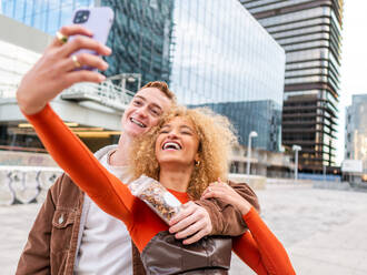 Delighted young multiracial couple in stylish wear cuddling and smiling while taking selfie on mobile phone standing near contemporary skyscrapers on city street - ADSF42722