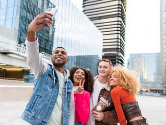 Optimistische junge multiethnische männliche und weibliche beste Freunde in stilvoller Freizeitkleidung, die lachen und lächeln, während sie ein Selbstporträt mit einem Smartphone auf einer Straße in der Nähe moderner Wolkenkratzer aufnehmen - ADSF42723