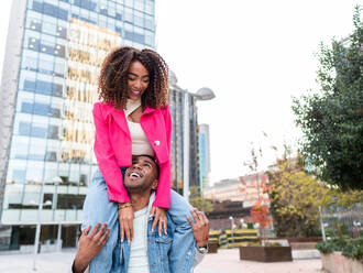 Cheerful young ethnic male millennial in casual clothes smiling happily while giving shoulder ride to delighted girlfriend against modern buildings on street - ADSF42726