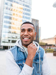 Confident cheerful young Hispanic male millennial in casual hooded jacket standing touching chin on street against modern buildings and looking away - ADSF42727