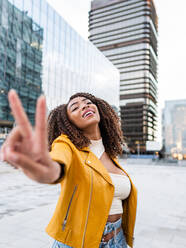 Delighted young ethnic female millennial with curly Afro hair in trendy crop top and yellow leather jacket jacket smiling and showing two fingers gesture standing on street against modern skyscrapers - ADSF42728
