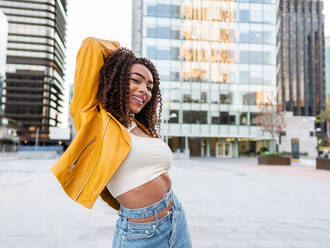 Selbstbewusste junge Afroamerikanerin mit langen dunklen lockigen Haaren in stilvollem Outfit, die fröhlich lächelt und in die Kamera schaut, während sie auf einer Straße in der Stadt steht und den Arm ausstreckt - ADSF42729