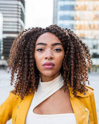 Confident young African American female model with curly dark hair and nose piercing in stylish yellow leather jacket standing on street near modern buildings and looking at camera - ADSF42730