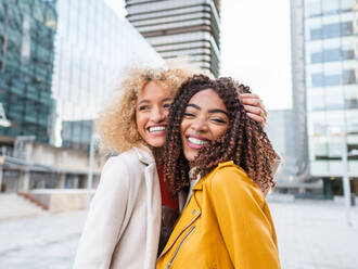 Seitenansicht von fröhlichen jungen stilvollen multirassischen Freundinnen mit lockigem Haar lächelnd beim Stehen umarmt einander auf Stadtstraße Blick auf die Kamera in der Nähe von zeitgenössischen Gebäuden - ADSF42731