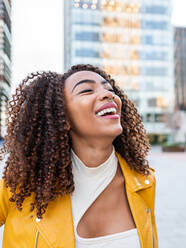 Self assured cheerful young African American female millennial with long dark curly hair in stylish outfit smiling brightly with eyes closed while standing on city street - ADSF42732