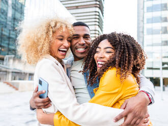 Delighted young ethnic guy with dark hair in casual clothes smiling while embracing cheerful diverse female friends standing on city street near modern buildings - ADSF42737