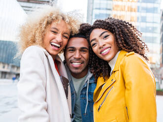 Delighted young ethnic guy with dark hair in casual clothes smiling while embracing cheerful diverse female friends standing on city street near modern buildings looking at camera - ADSF42738