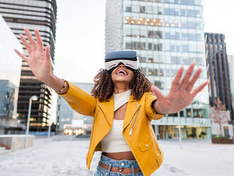Delighted young African American woman in stylish yellow jacket smiling while standing on city street experiencing virtual reality in modern headset - ADSF42740