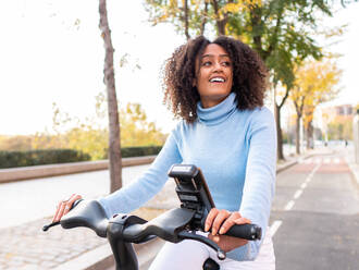 Fröhliche hispanische Frau in legerer Kleidung mit Afrofrisur fährt mit einem Elektrofahrrad durch eine Stadtgasse im Park und schaut weg - ADSF42742