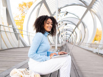 Seitenansicht von konzentrierten lächelnden ethnischen weiblichen Freiberufler in casual Outfit sitzt auf einer Holzbank in der modernen Stadt Brücke und arbeiten aus der Ferne über Laptop auf warmen Herbsttag Blick in die Kamera - ADSF42747