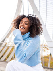 From below of cheerful young dreamy Hispanic female in warm sweater sitting on bench in modern bridge leaning on hand while looking at camera - ADSF42750