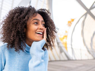 From below of cheerful young dreamy Hispanic female in warm sweater sitting on bench in modern bridge leaning on hand while looking away - ADSF42752