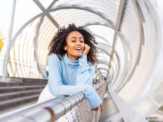 Lächelnde hispanische Frau in blauem Rollkragenpullover steht auf einer Brücke und stützt sich auf ein Metallgeländer, während sie an einem windigen Tag in die Kamera schaut - ADSF42753