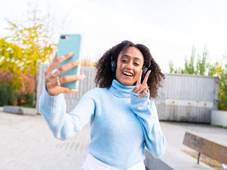 Positive Hispanic female with Afro hairstyle in blue turtleneck and headphones standing on road in park while listening to music and taking selfie on smartphone - ADSF42756