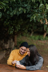 Hispanic man in outerwear pointing at smartphone screen for glad girlfriend while sitting at table on weekend day at park - ADSF42785