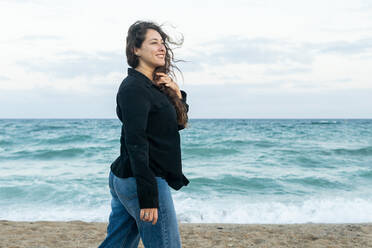 Side view of cheerful female in casual apparel standing near waving sea under cloudy sky on sandy coast smiling - ADSF42794