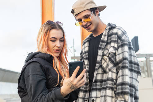 Niedriger Winkel eines jungen Mannes und einer Frau in modischer Kleidung, die ein Video auf ihrem Mobiltelefon ansehen, während sie gemeinsam Zeit auf einer Straße in der Stadt verbringen - ADSF42798