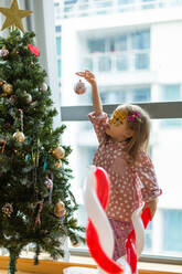 Seitenansicht eines bezaubernden Mädchens im rosa Schlafanzug mit Ballon-Zuckerstange, das sich zum Weihnachtsbaum streckt, um eine Weihnachtskugel aufzuhängen, während es in der Nähe des Fensters in einem hellen Raum zu Hause steht - ADSF42810