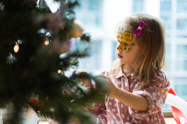 Side view of adorable girl with face painted with giraffe in pink pajama decorating Christmas tree with bauble while standing near window in light room at home - ADSF42811