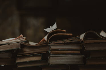 Kleines Papierschiff auf einem Stapel alter Bücher mit gebundenen Einbänden, die in einem dunklen Raum gestapelt sind - ADSF42815