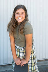 Positive girl in checkered skirt and t shirt standing against striped wall with skateboard - ADSF42824