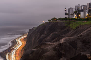 Long exposure shot of Miraflores boardwalk and curvy road with trails of light located near cliff with lighthouse and apartment buildings on shore of sea in cloudy evening in Lima, Peru - ADSF42840