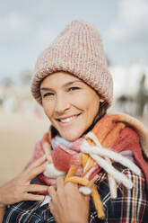Smiling woman wearing scarf and knit hat at beach - JOSEF16166