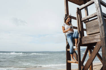 Frau mit Smartphone auf Rettungsschwimmer-Hütte am Strand sitzend - JOSEF16167