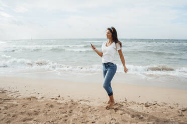 Woman using smart phone walking on sand at beach - JOSEF16238