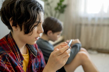 Boy playing video games on smart phone with brother sitting in background - ANAF00948