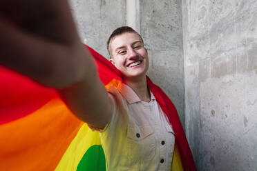 Fröhliche nicht-binäre Person, die ein Selfie mit Regenbogenflagge macht - ASGF03267