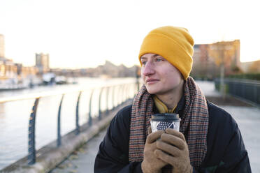 Smiling non-binary person with coffee cup standing at footpath - ASGF03285