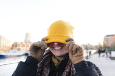 Happy non-binary person covering face with knit hat - ASGF03287