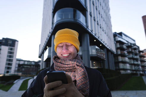 Happy non-binary person using smart phone in front of building - ASGF03288