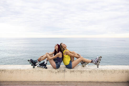 Frauen auf Inline-Skates sitzen Rücken an Rücken an der Promenade am Meer - WPEF07143