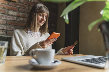 Smiling freelancer with credit card using smart phone at cafe - OSF01357