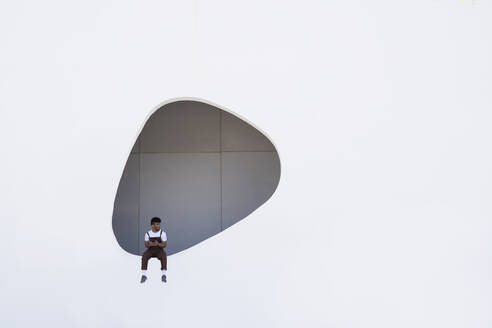 Junger Mann sitzt auf einer Mauer mit einem Loch im Gebäude - JCZF01140