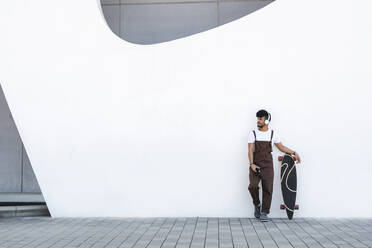 Young man wearing headphones standing by skateboard in front of wall - JCZF01151