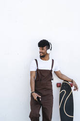 Smiling young man standing by skateboard in front of white wall - JCZF01152